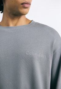 Šedá pletená mikina s kulatým výstřihem a vyraženým logem "Calvin Klein" na levé straně hrudi. Hladká struktura, ležérní design.