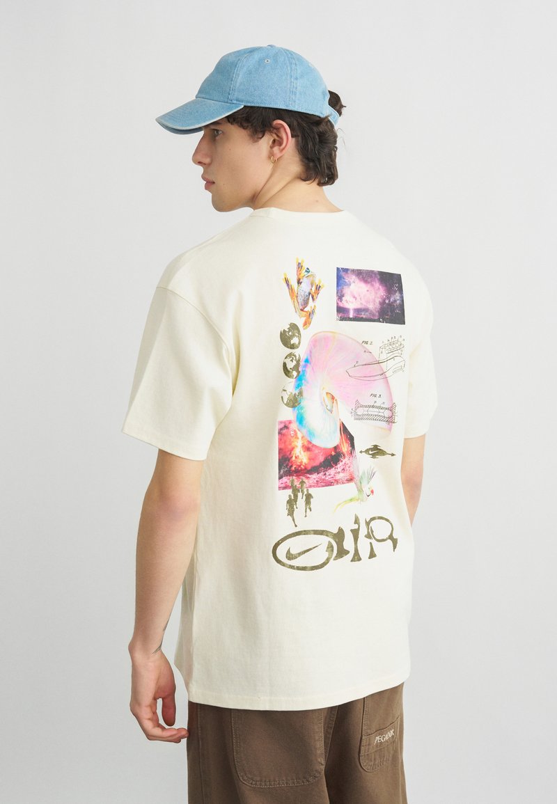 Ung mann i blå caps og beige t-skjorte med grafiske, abstrakte design og kosmisk bilde, stående foran en ensfarget lys bakgrunn.