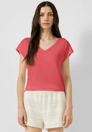 Woman with long brown hair wears coral V-neck short-sleeve top and white textured shorts, standing against a plain light background.