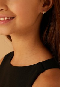 Gros plan d'une femme souriante portant une petite boucle d'oreille en or en forme de papillon et un haut sans manches noir, sur fond beige.