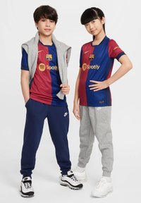 Djecak i djevojcica u sportskim odijelima: jedan u plavoj i bordo FC Barcelona majici, a drugi u sivoj trenerici. Oboje nose tenisice.