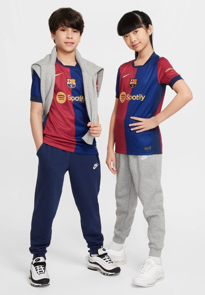 Djecak i djevojcica u sportskim odijelima: jedan u plavoj i bordo FC Barcelona majici, a drugi u sivoj trenerici. Oboje nose tenisice.