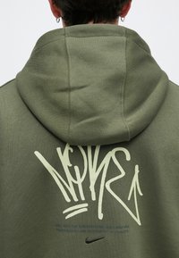 Ελιόχρωμο φούτερ με μεγάλη λευκή γραφιστική σχεδίαση και κυκλικό λογότυπο στην πλάτη. Η υφή του υφάσματος φαίνεται μαλακή, με άνετη κουκούλα.