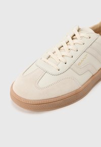 Κρεμ αθλητικά παπούτσια από δέρμα και σουέντ υλικά, με κορδόνια στο ίδιο χρώμα, σόλα από καουτσούκ με υφή και διακριτικό λογότυπο στο πλάι.