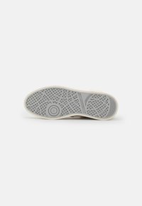 Rubberen sneakerzool in lichtgrijs met een gestructureerd patroon, voorzien van een cirkelvormig logo en maatindicatie. Strakke rand in crème kleur.