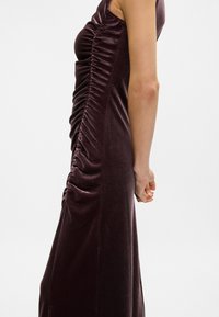 Μωβ βελούδινο φόρεμα με πλαϊνές ρυτίδες, χωρίς μανίκια και εφαρμοστή σιλουέτα. Υφασμα με υφή και απαλό γυάλισμα.