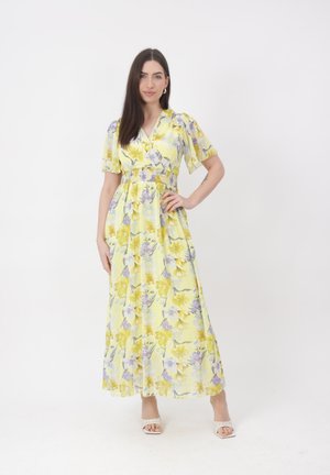Kobieta o długich ciemnych włosach, ubrana w żółtą, kwiecistą sukienkę maxi z krótkimi rękawami i białe sandały na obcasie, stojąca na tle białego tła.