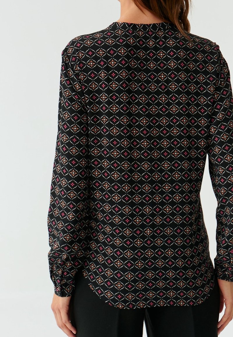 Femme portant un chemisier noir à manches longues avec un motif géométrique rose et beige, vue de dos sur un fond uni.