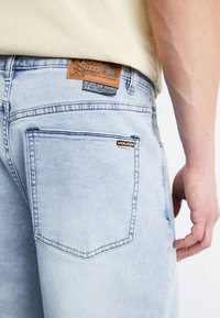 Jeans de mezclilla azul claro con un parche de cuero marrón que presenta la marca. Incluye un pequeño logo negro encima del bolsillo trasero.