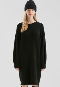 Jonge vrouw met een zwarte gebreide beanie en een oversized lange mouw zwarte gestructureerde truitjurk, staand tegen een effen lichte achtergrond.