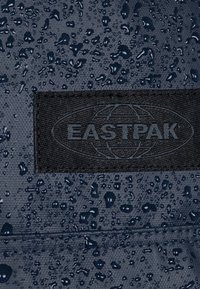 Tissu imperméable bleu marine avec des gouttes d'eau, présentant une surface texturée. Étiquette logo Eastpak noire appliquée pour identification.