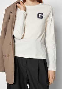 Weißes Langarm-Chemise aus Baumwolle mit einem marineblauen Grafik-"G"-Logo auf der Brust, kombiniert mit einem braunen Blazer und schwarzen Hosen.