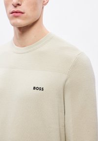 Bézs színű kötött pulóver texturált mintával, kerek nyakkivágással és fekete "BOSS" logóval a bal mellrészen. Simogató anyag bordás részletekkel.