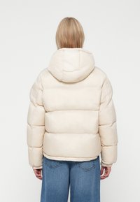Puffjacka i ljusbeige, med huva, quiltat mönster och ribbade ärmslut. Mjuk, texturerad tyg med en voluminös silhuett.