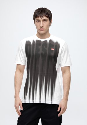 Jeune homme aux cheveux foncés portant un T-shirt blanc avec des rayures verticales noires et un petit logo rouge Diesel sur un fond uni.