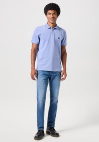 Hombre con cabello rizado que lleva una camisa polo azul claro, jeans azules y zapatos negros, de pie y sonriendo frente a un fondo sencillo.
