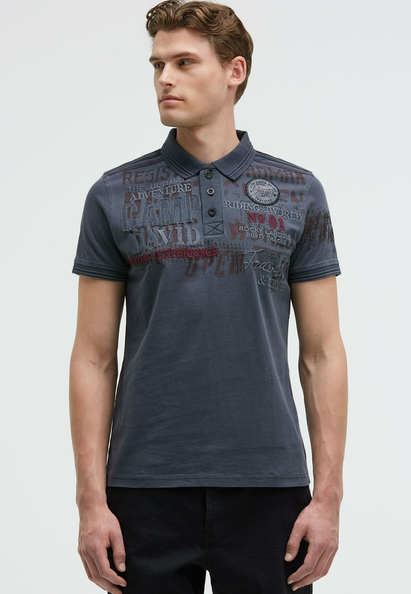 Dark blue polo shirt made of cotton, featuring a collar, three-button placket, and mixed text graphics in red and gray tones.
