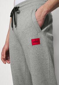 Pantalones deportivos grises hechos de un material suave, con cintura elástica y cordón negro. Presenta un parche rectangular rojo con la inscripción "HUGO".