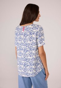 Kurze Ärmelbluse mit weißem Grund und blauem Blumenmuster, mit einem Rückenschlitz-Detail und pinkem Akzent. Weicher Stoff, entspannter Schnitt.