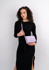 Borsa a tracolla lilla con texture liscia, forma rettangolare e tracolla. Indossata con un vestito nero a coste con spacco laterale.