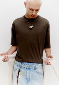 Tmavě hnědé oversized tričko s krátkým rukávem, doplněné o světle modré džíny s efektem opotřebení. Doplňky zahrnují stříbrný náhrdelník a barevné přívěsky.