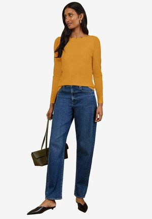 Žena nosiaci horčicovo žltý top s dlhým rukávom, modré džínsy s vysokým pásom, čierne špicaté topánky a nesúca olivovo zelenú kabelku.