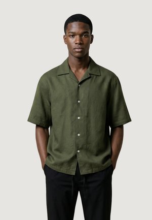 Giovane uomo in piedi con le mani nelle tasche, indossa una camicia verde oliva a maniche corte con bottoni e pantaloni neri, davanti a uno sfondo semplice.