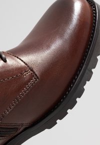 Botte en cuir marron avec une finition brillante, semelle noire robuste, détails cousus, et design à bout rond, mettant l'accent sur la durabilité et le confort.