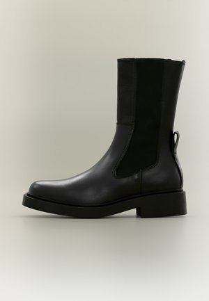 Schwarzer Leder-Stiefelette mit elastischen Seitenteilen, einer Zuglasche hinten und niedrigem Blockabsatz auf einfarbig hellem Hintergrund.