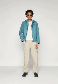 Gaiši zila ūdensnecaurlaidīga jaka ar kapuci, uzvilkta virs baltas krekliņas. Kombinēta ar beidžas krāsas kravas biksēm un melniem apaviem, kā arī ar saulesbrillēm.