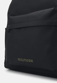 Musta reppu, joka on valmistettu sileästä materiaalista, esillä edessä oleva tasku ja kohokuvioitu "HILFIGER"-logo oliivinvihreänä.