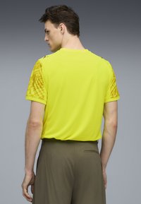 Żółta koszulka sportowa z krótkim rękawem z teksturowanym wzorem na rękawach i szczegółem logo. Posiada okrągły dekolt i luźny krój.