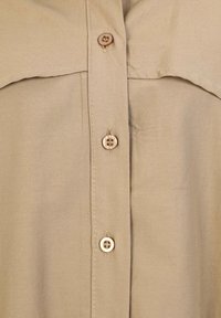 Beige overhemd met knopen en een zachte textuur, met een puntige kraag, dubbele borstzakken en bruine knopen. Eenvoudig en neutraal ontwerp.
