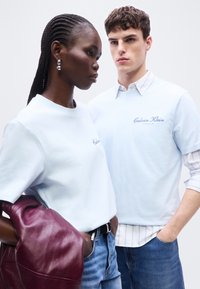 Γυναίκα και άντρας με ανοιχτό γαλάζιο πουκάμισο Calvin Klein, η γυναίκα κρατάει ένα δερμάτινο μπουφάν μπορντό, και οι δύο στέκονται μπροστά σε απλό φόντο.