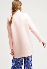 Chaqueta de manga larga rosa claro con un diseño simple y un corte relajado, combinada con pantalones azules de estampado floral para crear contraste.