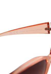 Solglasögon med halvgenomskinlig orange båge, mörka linser och metallgångjärn. Märkningar på insidan av skänkeln visar "Tillverkad i Kina" och ett modellnummer.