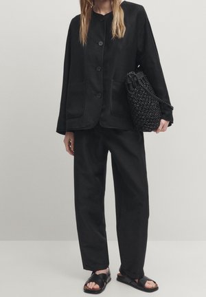Woman wearing loose black buttoned jacket and pants, holding a textured black woven bag, and black sandals on light background.