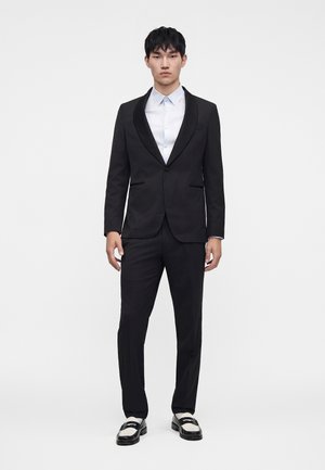 Giovane uomo in piedi di fronte, indossa uno smoking nero con rever in satin, camicia bianca e mocassini bianchi e neri su sfondo bianco uniforme.
