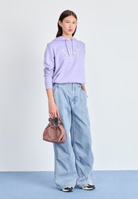 Haut à capuche lilas avec logo blanc, jean large bleu clair, et un petit sac marron avec une anse froncée. Des baskets noir et blanc complètent le look.