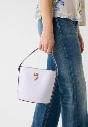 Frau trägt blaue Jeans und eine geblümte Bluse, hält eine weiße Eimerhandtasche mit kurzem Griff und goldfarbenem Verschluss.