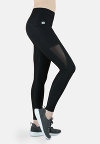 Hoch taillierte schwarze Leggings mit seitlichen Netzeinsätzen, genähten Details und einem Logo. Kombiniert mit grauen Sportschuhen auf einem hellen Hintergrund.