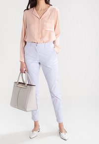 Ljusrosa satinblus med en ficka, tillsammans med ljusblåbyxor. Modellen håller en grå strukturerad handväska och bär vita spetsiga skor.