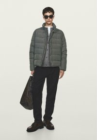 Gepolsterte olivgrüne Jacke, grauer Strickpullover, schwarze Hose, Ledertasche und dunkle Schuhe. Glatte Oberfläche mit Steppdetail auf der Jacke.