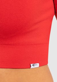 Rode crop top met een gladde textuur, voorzien van een ribbelrand en een klein logo label aan de zijkant. Strak design ideaal voor activewear.