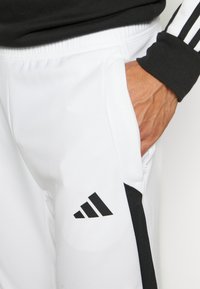 Weiße Sporthose mit schwarzen Akzenten und dem Adidas-Logo. Der Stoff wirkt glatt und hat einen elastischen Bund sowie Taschen.
