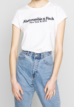 Kobieta ubrana w białą koszulkę Abercrombie & Fitch i jasnoniebieskie jeansy z wysokim stanem, stojąca z rękami swobodnie opuszczonymi wzdłuż ciała.