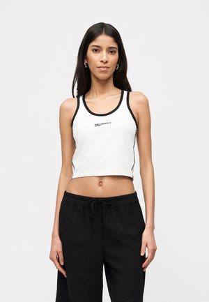 Jeune femme portant un débardeur court blanc avec bordure noire et texte, associé à un pantalon noir ample avec cordon de serrage, debout devant un fond uni.