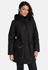 Vrouw in een zwarte gewatteerde winterjas met capuchon, met voorzakken en een ritssluiting, staande tegen een effen lichte achtergrond.