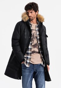 Parka nera con cappuccio foderato in pelliccia, dotata di zip frontale, cordini regolabili e numerose tasche, indossata sopra una camicia a quadri e jeans.