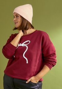 Sudadera color burdeos con gráfico de lazo blanco, cuello redondo y puños de canalé, combinada con un gorro de punto con canalé en rosa claro. Fondo verde.